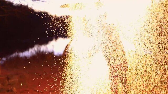 Young male performing dance with charcoal staff and flame in sparkle like, fire pois near lake at night during fire jamming in forest near lake
