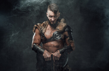 Obraz na płótnie Canvas Brutal nordic heathen dressed in light armour posing in dark smokey background with huge axe and staring at camera.