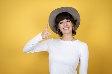 Young caucasian woman wearing hat over isolated yellow background smiling and doing the ok signal with her thumb