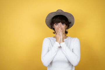 Young caucasian woman wearing hat over isolated yellow background with her hands over her mouth and surprised