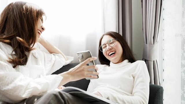 Daily Holiday Life Of A Single Middle Aged Woman In The Living Room Of The House. Happy Asian Woman Lying On Sofa Play Smartphone With Friend Sitting And Reading Book Together.