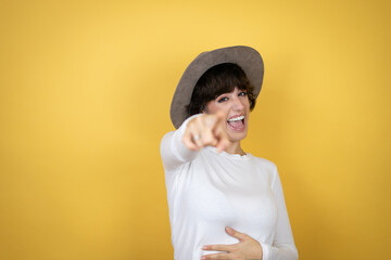 Young caucasian woman wearing hat over isolated yellow background laughing at you, pointing finger to the camera with hand over body, shame expression