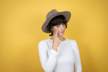 Young caucasian woman wearing hat over isolated yellow background Pointing to the eye watching you gesture, suspicious expression