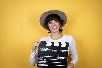 Young caucasian woman wearing hat over isolated yellow background holding clapperboard very happy...