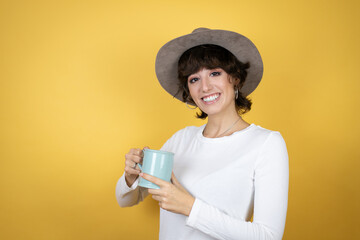 Young caucasian woman wearing hat over isolated yellow background enjoying and drinking a cup of...