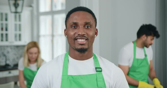 Attractive smiling experienced black-skinned worker of cleaning service poses on camera on the background of male and female colleagues which cleaning in client's house