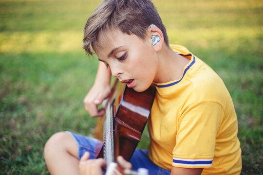 Hard of hearing preteen boy playing guitar and singing. Child with hearing aids in ears playing music and singing song in a park. Hobby art activity for children kids. Authentic childhood moment.