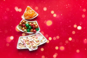 on a red background there is a tray in the form of a Christmas tree. sweets in the tray: marshmallows, round candies in foil and tangerine slices