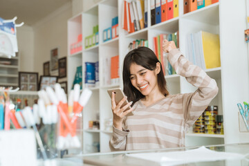 Portrait cheerful young girl entrepreneur happy to see orders via cellphone working in a stationery shop