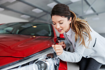 Smiling female car seller in suit wiping car with her sleeve while standing in exclusive car salon.