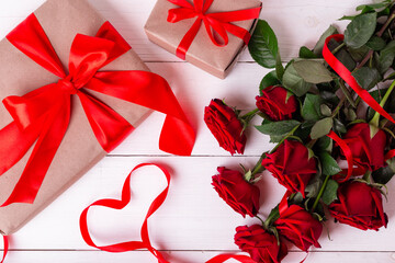 St. Valentines Day concept. Red ribbon shaped as heart, bouquet of roses and gift boxes wrapped in kraft papper on white wooden table.