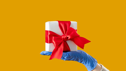 Holiday gift. Happy valentine day. Covid-19 restriction. Hand in protective gloves holding present box taped red ribbon isolated on yellow copy space. Advertising background.