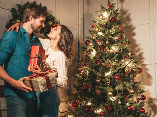 Obraz na płótnie Canvas Smiling beautiful woman and her handsome boyfriend. Happy cheerful family posing in the interior near Christmas tree. Models hugging. Love, x-mas concept. They holding gift boxes for each other