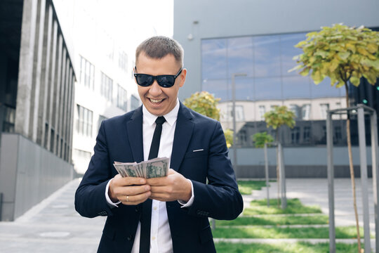 Rich man in stylish suit wearing sunglasses counting money and walking near office building. Looking successful, confident