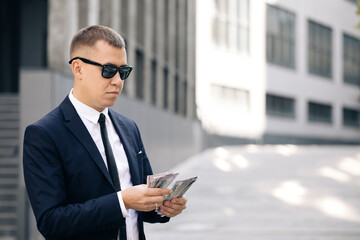 Portrait of handsome caucasian young businessman in sunglasses counting money while standing outdoors. Joyful male with many dollars. Luxury life. Money concept