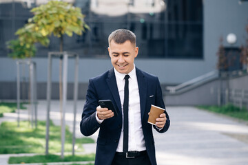 Stylish Businessman Exiting from the Modern Office. Using his Smartphone. Sliding on Mobiles Screen. Drinking Tasty Coffee. Looking Successful, Confident. Holding Coffee Cup on Summer Day