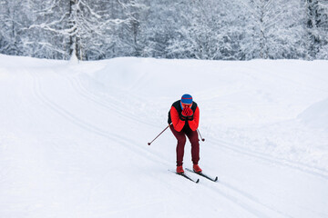 A skier in a red tracksuit, blue balaclava, and safety glasses riding fast downhill, covering his face with his hands from the cold wind with a snow-covered forest background on a cloudy day