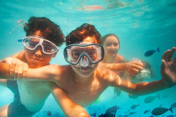 Two happy boys in scuba mask dive underwater with friends in sea among fish smile and play