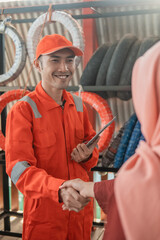 male mechanic in a wearpack uniform holding a digital tablet shake hands with a female customer in a veil at a spare part workshop