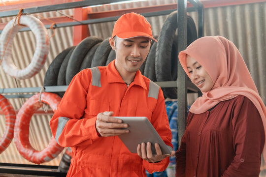 a male mechanic in a wearpack uniform and a female customer in a veil using a digital tablet to view the catalog at a spare part workshop