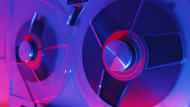 Reel to reel tape recorder playing ultra red blue neon light. Rotating vintage music player close up. Retro tape. Spinning reels metallic color with red disc. Popular Disco Trends 60s, 70s, 80s, 90s.
