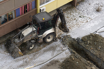 An excavator digs a trench in winter. The excavator is working next to a residential building. Excavator tracks in the snow. The excavator bucket is raised.