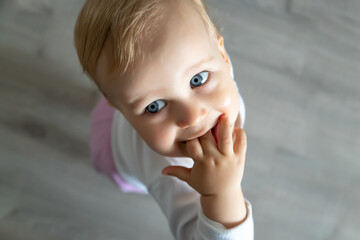Cute adorable infant blond happy little toddler boy stand in home room looking above and sucking hand in mouth against floor background. Child virus and sick runny nose. Kid healthcare concept