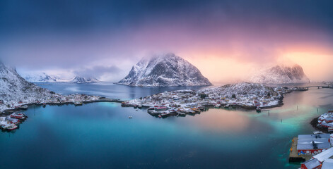 Aerial view of blue sea, snowy mountains, high rocks, village with buildings, rorbu, colorful sky, reflection in azure water. Reine at sunset in winter. Top view of Lofoten islands, Norway. Landscape