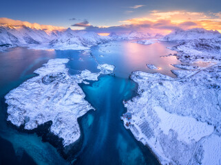 Aerial view of Lofoten islands in winter at sunset in Norway. Landscape with blue sea, snowy mountains, rocks and islands, road, bridge, village, buildings, colorful sky with pink clouds. Top view