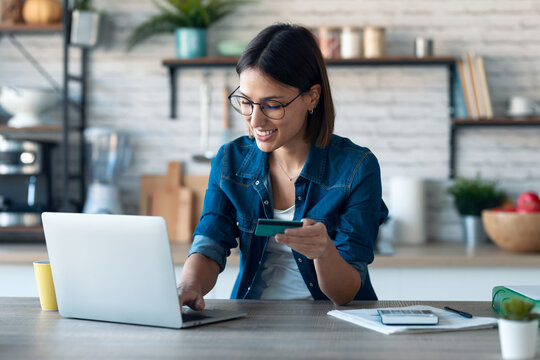 Cute young woman holding white credit card for shopping online with computer while sitting in the kitchen at home.