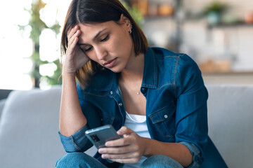 Worried young woman using her mobile phone while thinking about problems sitting on couch in the living room at home.