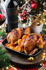 Holiday baked quail or chicken with dried apricots in pan on red napkin served with glasses, bottle of wine, candles, fir branches, festive decoration, garland. Christmas and New Year food, bokeh
