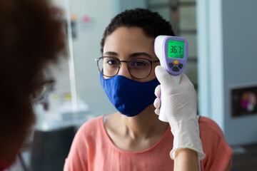 Mixed race woman wearing face mask checking temperature of female colleague