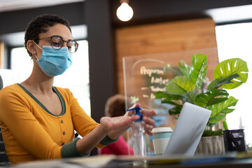 Mixed race woman wearing face mask sanitizing hands at desk in office
