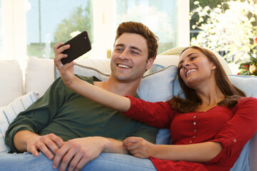 Caucasian couple taking selfie with smartphone sitting on couch smiling