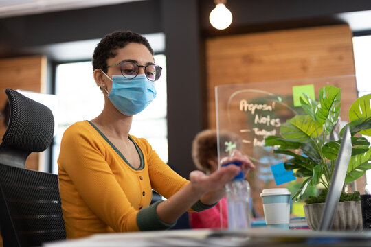 Mixed race woman wearing face mask sanitizing hands at desk in office - Powered by Adobe