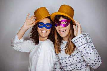 .Two beautiful women saying goodbye to the year 2020 in pajamas from home due to the pandemic caused by covid19. Festive and fun attitude wearing golden hat and colorful mask