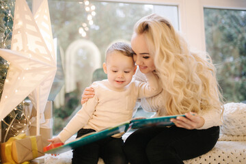 Happy little boy read book with mom on windowsill. Mom with her cute son spend time together on Christmas holiday. Decorated room