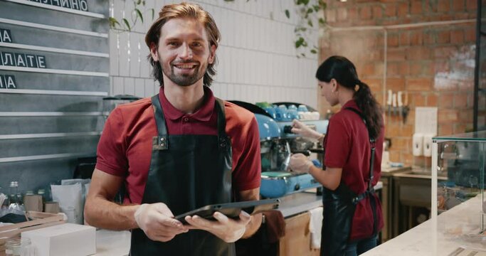 Caucasian hanfsome male barista employee wearing apron using digital tablet computer and smiling to camera. Occupation. Coffeeshop. Coffee house.