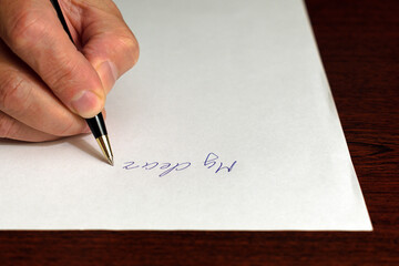 beginning of the letter on white sheet of paper. hand with ballpoint pen. focus in center of image. view out of side