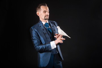 A man magician with a beard and mustache in a blue tailcoat holding a dove on a black background