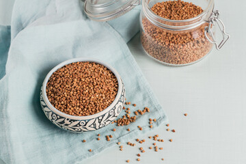 Bowl of dry raw buckwheat groats and glass jar on a light blue background. Cooking buckwheat porridge concept.