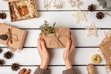 Flat lay of gift craft boxes, pine cones, cinnamon sticks, dried flowers and Christmas decorations on a wooden light background. Eco friendly christmas. Female hands are holding a gift.