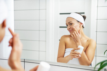 A young smiling woman wrapped in towels holding moisturizing cream and applying it on her face in bathroom.