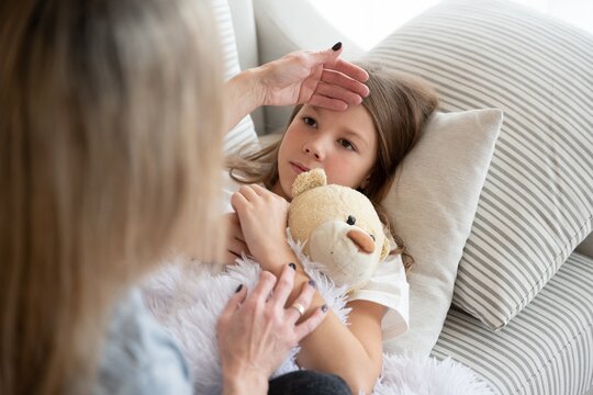 The sick girl is lying with the teddy bear. Mom is checking for a fever.
