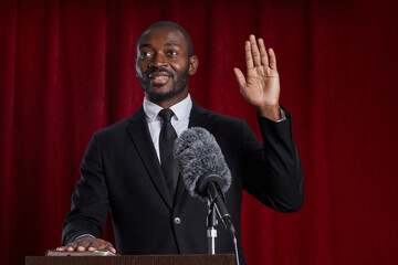 Waist up portrait of African-American man giving oath while standing at podium on stage against red...