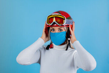 Close-up portrait of a girl skier wearing a light blu protective mask, helmet and glasses. A snowboarder on an light blu background. Covid19 coronavirus and the ski resort pandemic.