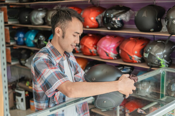 side view of male shop assistant stands cleaning a helmet with a cloth with helmet display rack background in a helmet shop