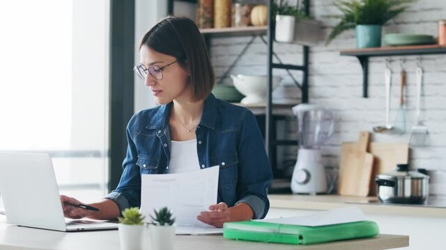 Video of pretty young business woman working with computer while consulting some invoices and documents in the kitchen at home.