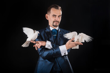A magician in a blue tailcoat holds two white doves in his hands on a black background